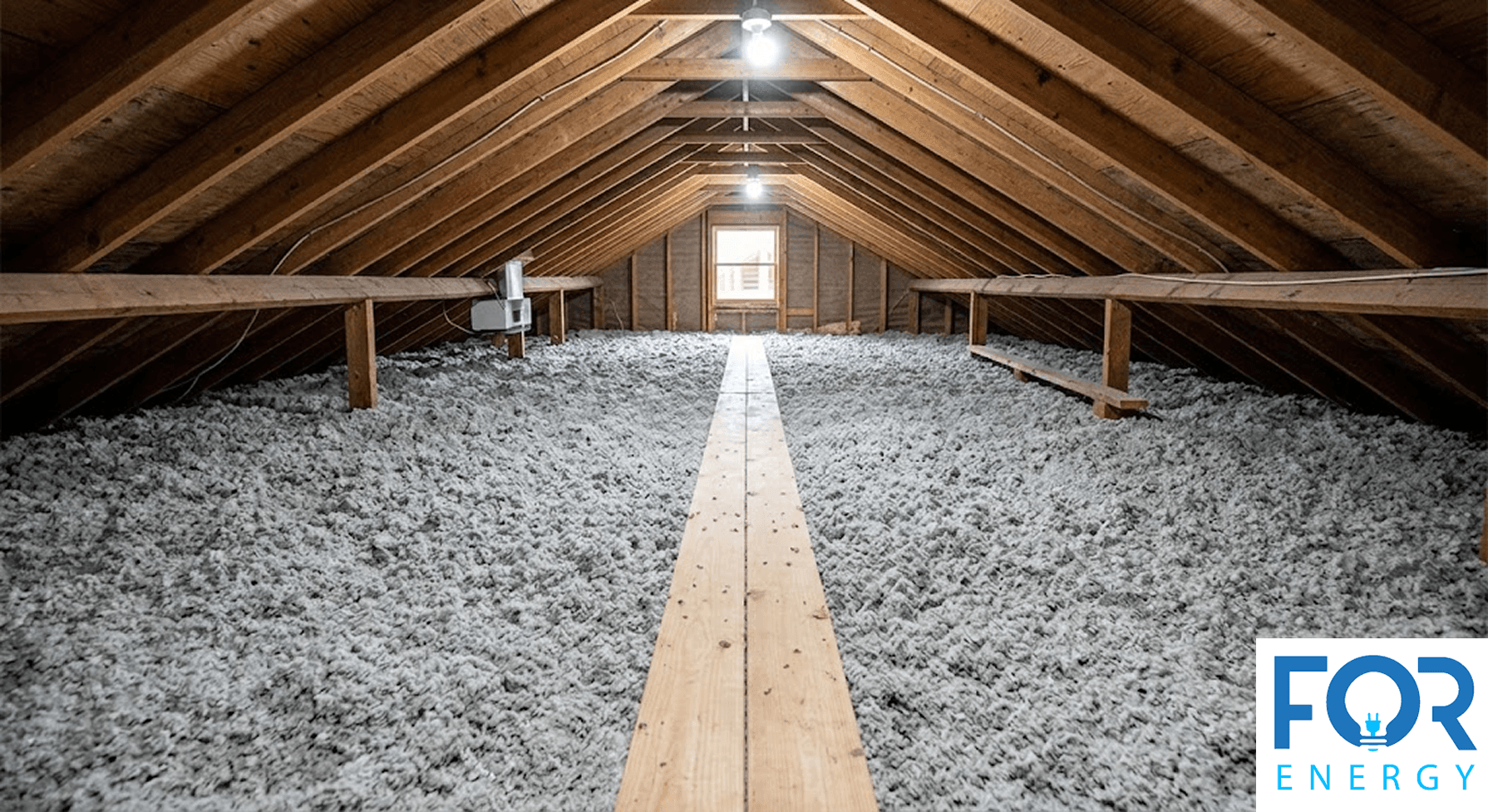 A wide-angle view of a professionally insulated attic featuring a thick, uniform layer of grey blown-in cellulose insulation. A long wooden walkway runs down the center of the space under a gabled roof with exposed timber rafters, lit by overhead work lights. The "FOR ENERGY" logo is in the bottom right corner.